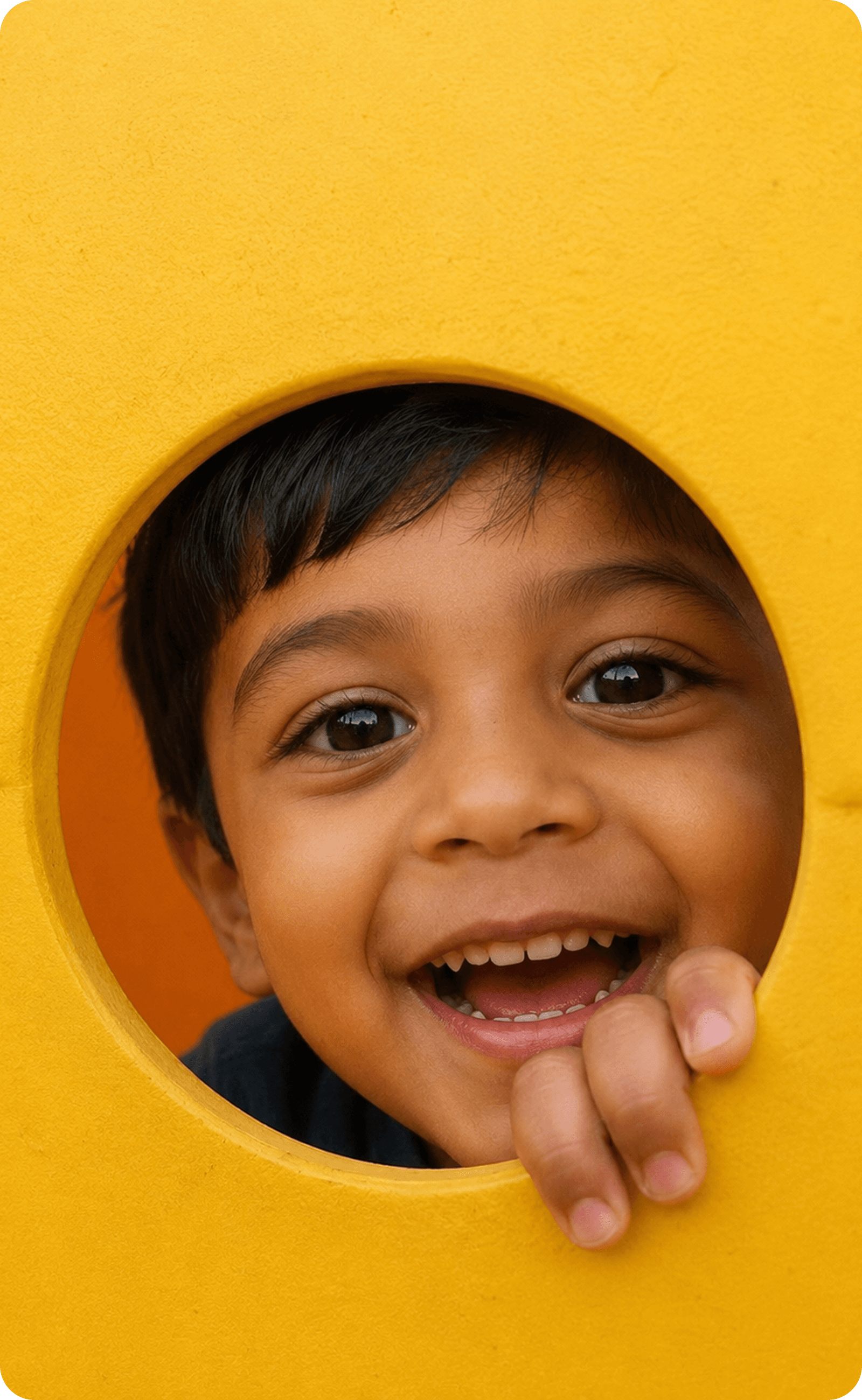 A boy laughing as he looks through a round hole in a yellow playground panel