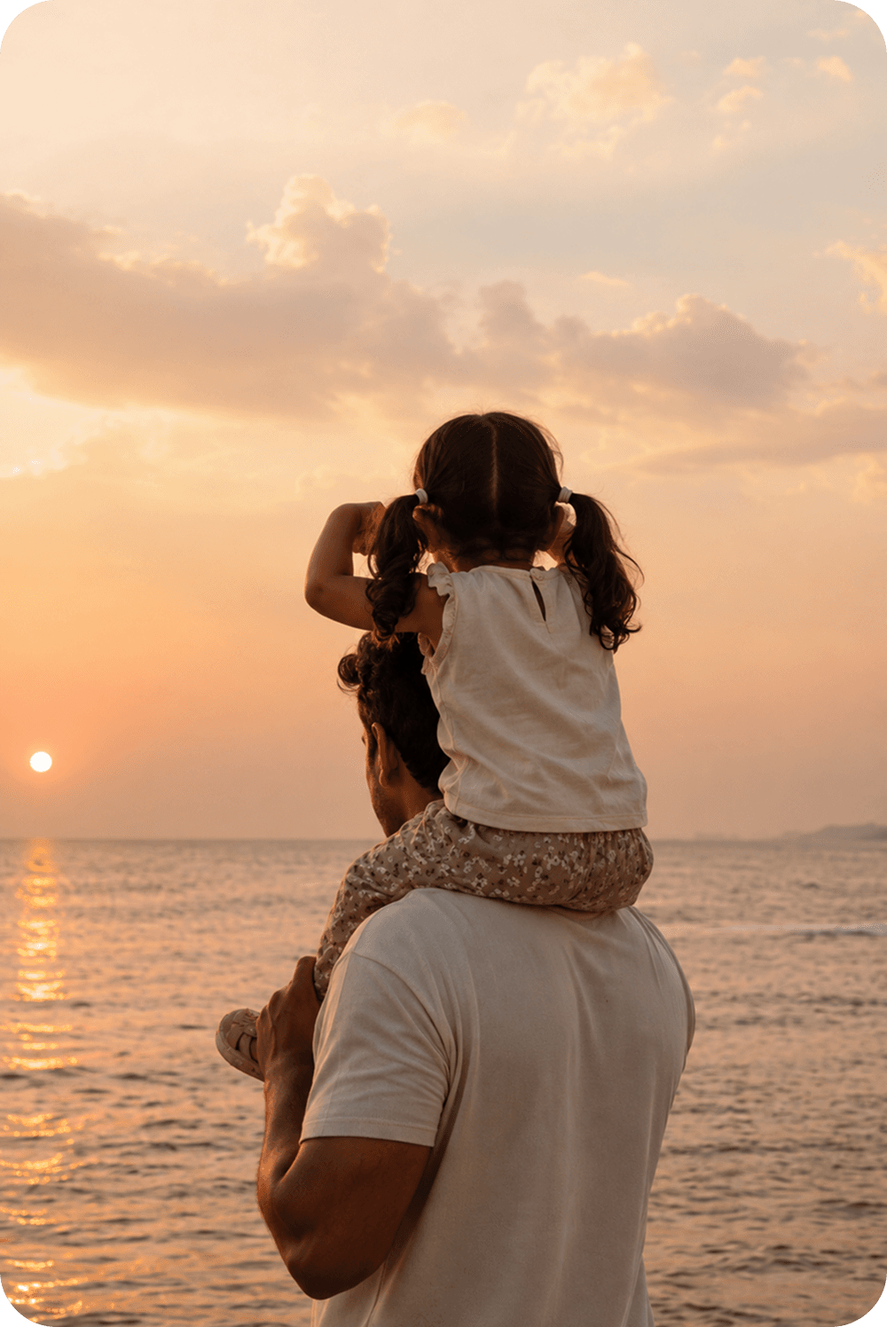 A father carrying his daughter on his shoulders, watching the sun set over the sea